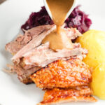 Onion gravy being poured over the meat from a beer roasted pork knuckle, served with potatoes and red cabbage.