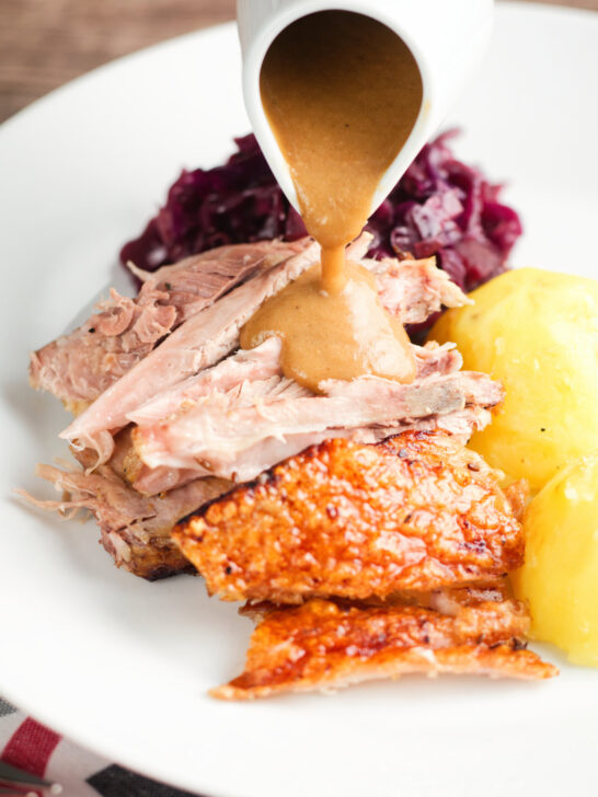 Onion gravy being poured over the meat from a beer roasted pork knuckle, served with potatoes and red cabbage.