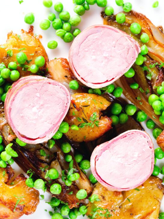 Overhead close-up bacon wrapped pork tenderloin, crushed roasted potatoes, braised chicory and peas.