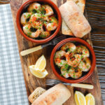 Overhead Spanish gambas (prawns) pil pil served with fresh parsley, bread and lemon wedges.