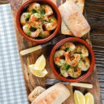 Overhead Spanish gambas (prawns) pil pil served with fresh parsley, bread and lemon wedges.