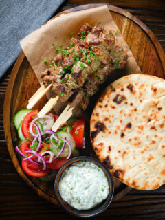 Overhead lamb souvlaki skewers served with tzatziki, salad and Greek flatbreads.