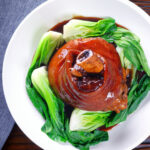 Overhead Chinese braised pork knuckle or shank served with steamed pak choi and braising juices.