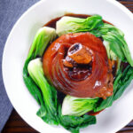 Overhead Chinese braised pork knuckle or shank served with steamed pak choi and braising juices.