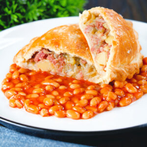 Homemade corned beef, onion and potato pasty cut open to show filling served with baked beans.