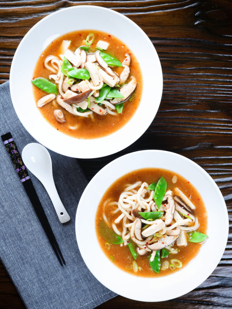 Two bowls (overhead) of vegan miso noodle soup with tofu, mangetout and shiitake mushrooms.