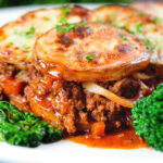 Close-up minced beef and onion hotpot with scalloped potatoes served on a plate with green veggies.