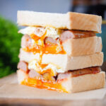 Close-up sausage and fried egg breakfast sandwich with a runny yolk served with coffee.