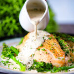 Creamy tarragon sauce being poured over chicken breast with celeriac mash and broccoli.