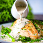 Creamy tarragon sauce being poured over chicken breast with celeriac mash and broccoli.