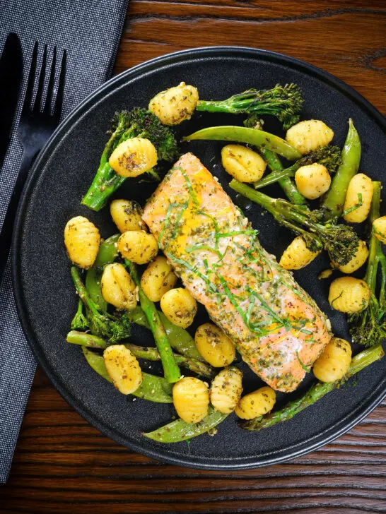 Overhead salmon fillet tray bake with pesto, green veggies and gnocchi served on a black plate.