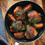 Overhead roasted honey and harissa chicken thighs with red onions cooked in a small frying pan.