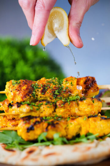 Lemon wedge being squeezed over Persian joojeh kabob (saffron chicken) with flatbread and rocket.