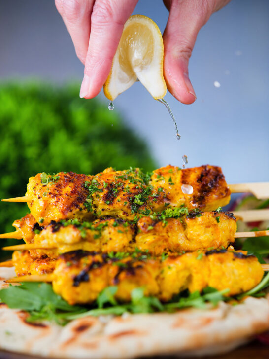 Lemon wedge being squeezed over Persian joojeh kabob (saffron chicken) with flatbread and rocket.