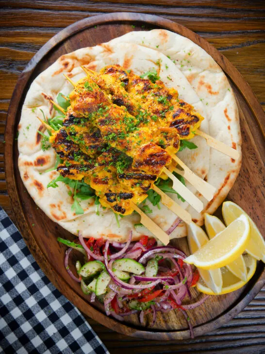 Overhead Persian joojeh kabob (saffron chicken skewers) with flatbread, Shirazi salad and lemon wedges.