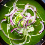 Close-up overhead palak paneer, Indian spinach curry garnished with red onion and cream.