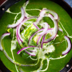 Close-up overhead palak paneer, Indian spinach curry garnished with red onion and cream.