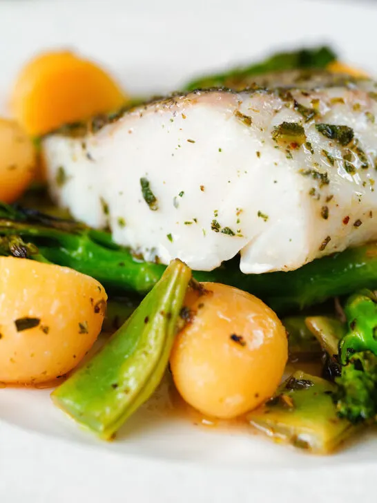 Close-up cod traybake with potatoes, broccoli and runner beans cooked in a herby olive oil.