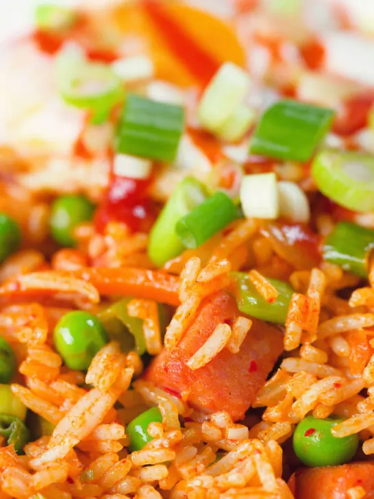 Close-up spicy Spam fried rice with kimchi and peas, served with a fried egg and Sriracha sauce.