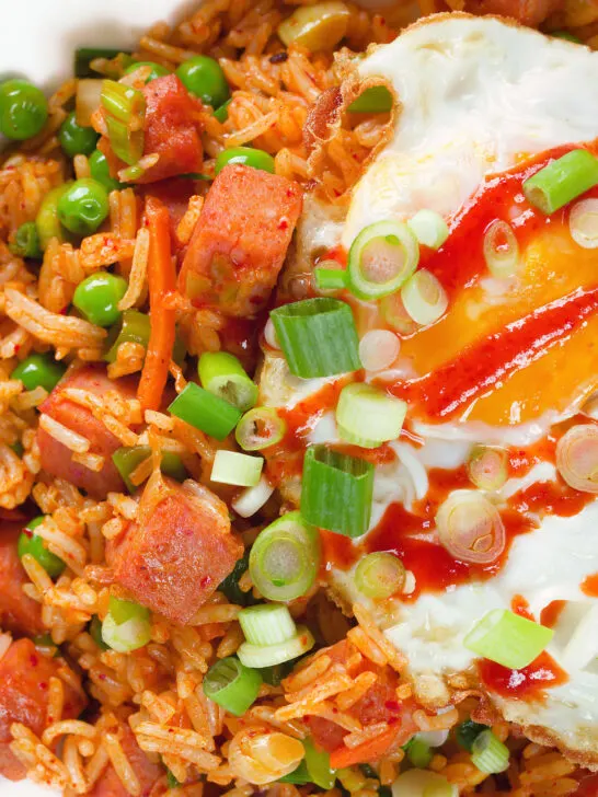 Close-up overhead spicy Spam fried rice with kimchi and peas, served with a fried egg and Sriracha sauce.