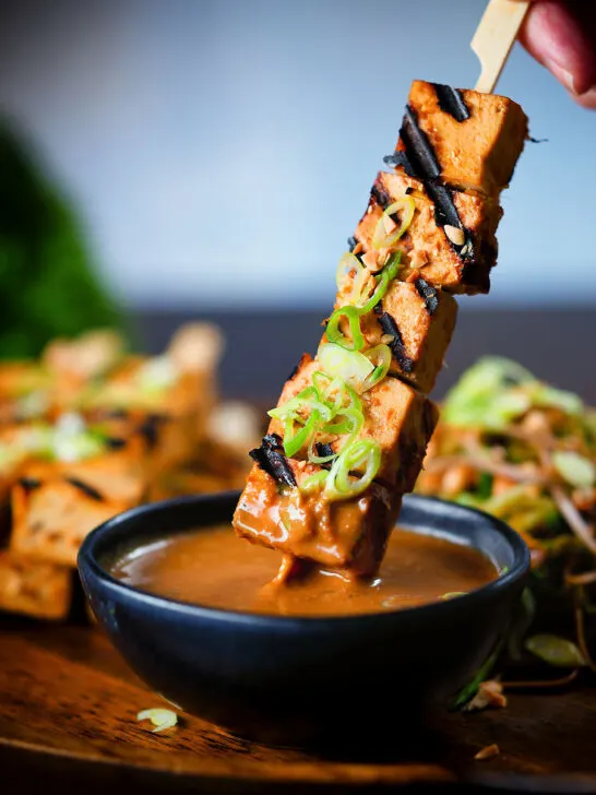 Tofu satay skewers being dipped into a homemade peanut dipping sauce.
