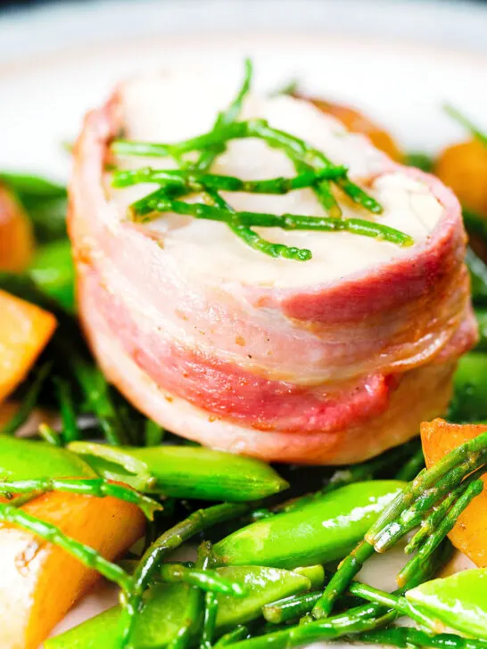Close-up bacon wrapped monkfish with potatoes, samphire and sugar snap peas in butter and cider.
