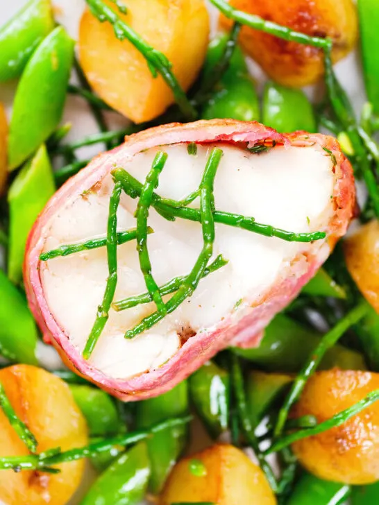 Close-up overhead bacon wrapped monkfish with potatoes, samphire and sugar snap peas in butter and cider.