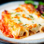Close-up baked three cheese, pork sausage, leek and ricotta stuffed cannelloni served with a side salad.