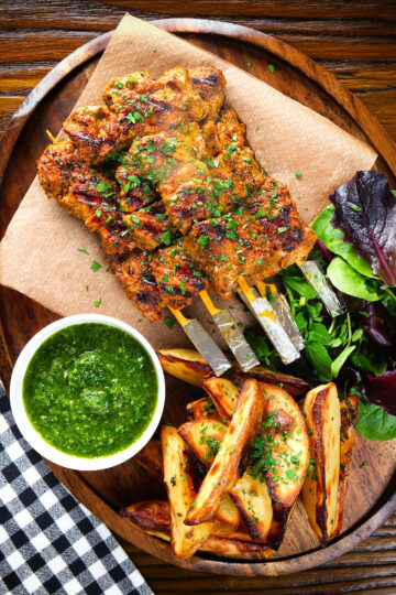 Overhead Spanish Pinchos morunos (pork skewers) served with a parsley salsa and potato wedges.
