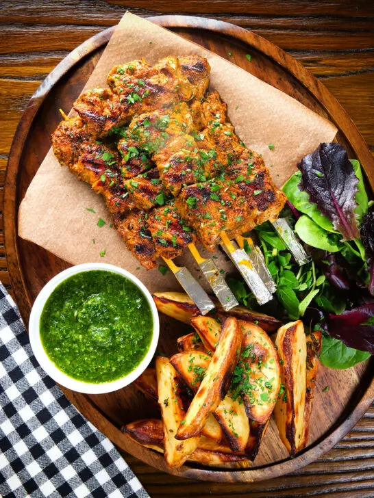Overhead Spanish Pinchos morunos (pork skewers) served with a parsley salsa and potato wedges.