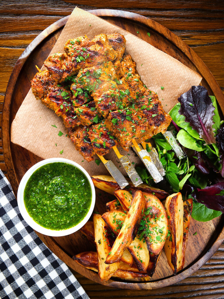 Overhead Spanish Pinchos morunos (pork skewers) served with a parsley salsa and potato wedges.
