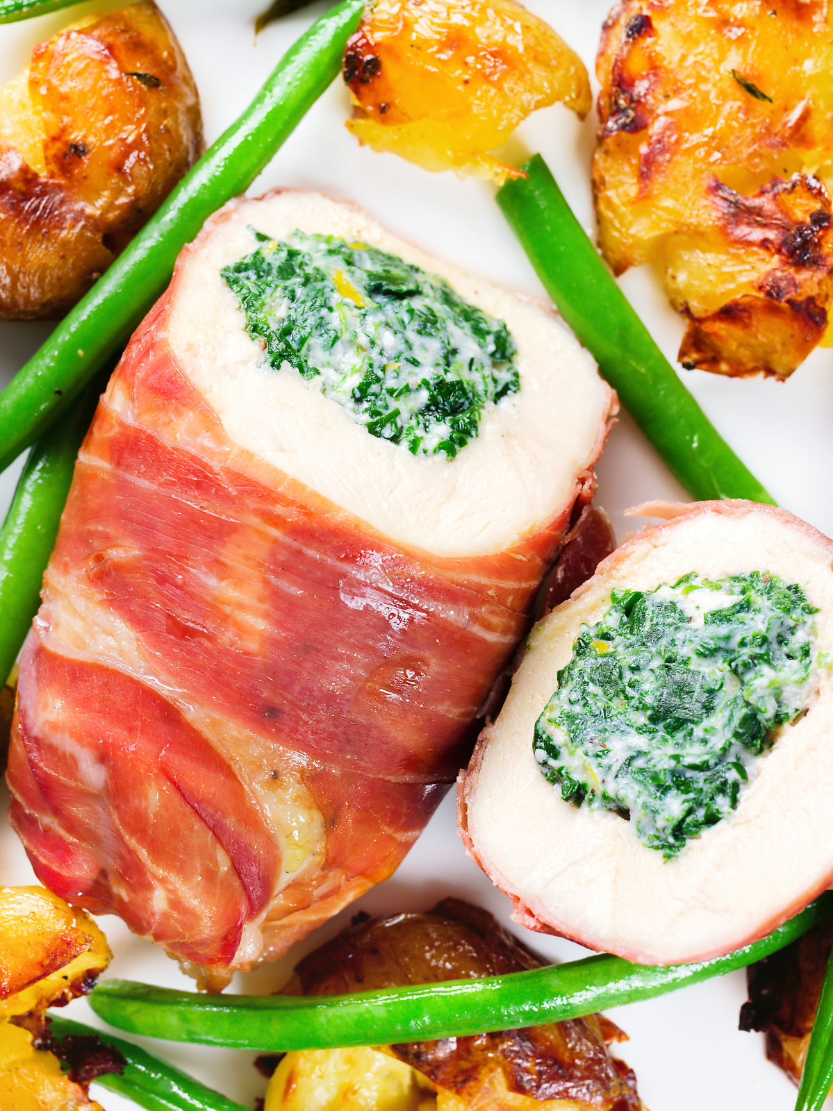 Close-up overhead chicken wrapped in Parma ham stuffed with spinach and ricotta served with roasted new potatoes and green beans.