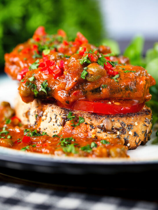Close-up tinned sardines on toast in tomato sauce with capers, fresh tomato, and chilli.