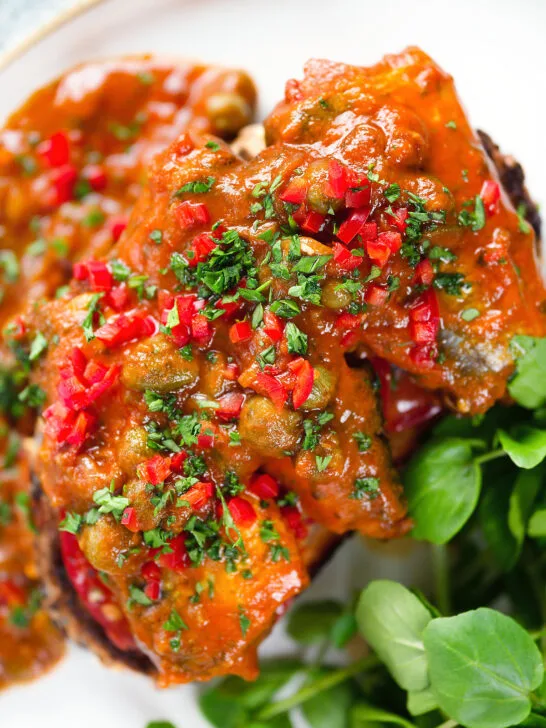 Close-up overhead tinned sardines on toast in tomato sauce with capers, fresh tomato, and chilli.