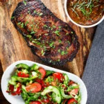 Overhead Thai Crying Tiger Steak (resting) with a spicy dipping sauce, and tomato and cucumber salad.