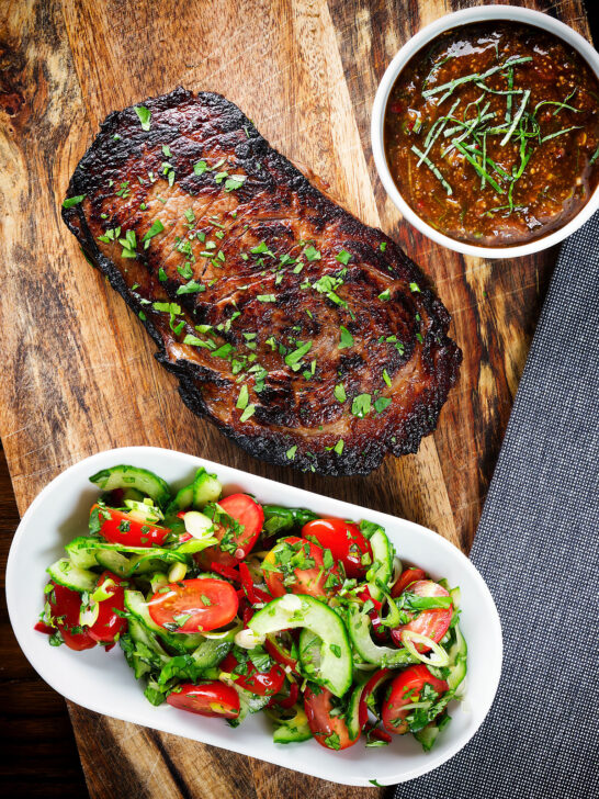 Overhead Thai Crying Tiger Steak (resting) with a spicy dipping sauce, and tomato and cucumber salad.