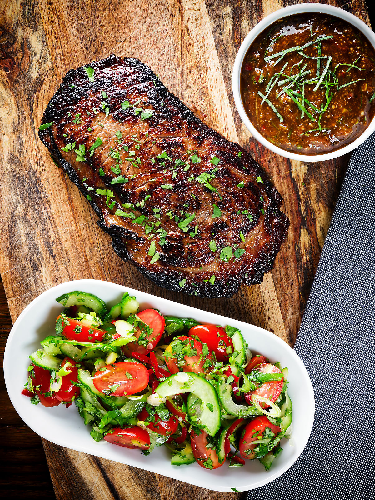 Overhead Thai Crying Tiger Steak (resting) with a spicy dipping sauce, and tomato and cucumber salad.