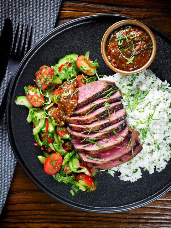 Overhead Thai Crying Tiger Steak with a spicy dipping sauce, rice, and tomato and cucumber salad.