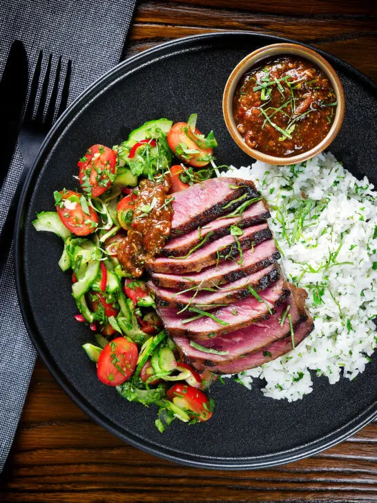 Overhead Thai Crying Tiger Steak with a spicy dipping sauce, rice, and tomato and cucumber salad.