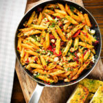 Overhead rose harissa, sun-dried tomato, pepper and feta cheese pasta in a pan with garlic bread.