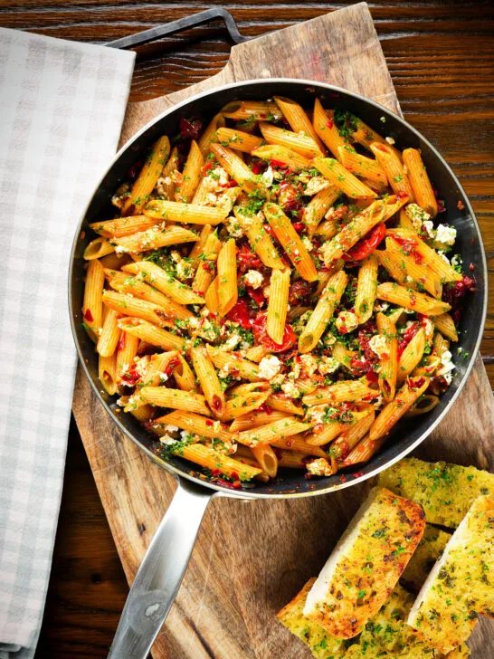 Overhead rose harissa, sun-dried tomato, pepper and feta cheese pasta in a pan with garlic bread.