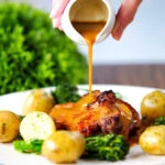 Cider gravy being poured over baked apple and bacon stuffed pork chops served with potatoes and broccoli.