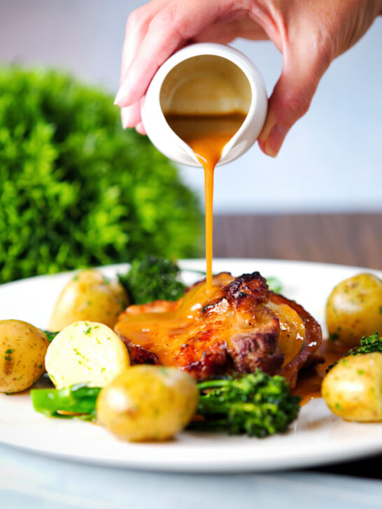 Cider gravy being poured over baked apple and bacon stuffed pork chops served with potatoes and broccoli.