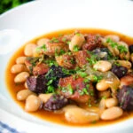 Pork and bean stew with black pudding and sage served in a white bowl.