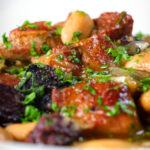 Close-up pork and bean stew with black pudding and sage served in a white bowl.