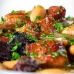 Close-up pork and bean stew with black pudding and sage served in a white bowl.