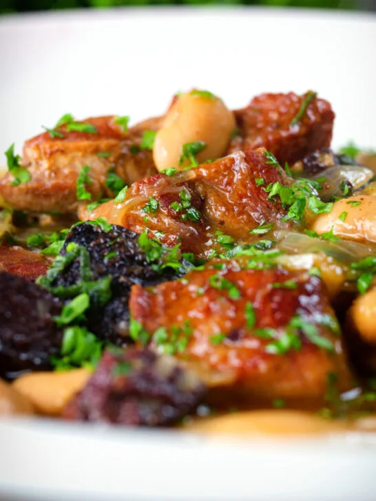 Close-up pork and bean stew with black pudding and sage served in a white bowl.
