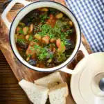 Overhead pork and bean stew with black pudding and sage served in its cooking pot with bread.