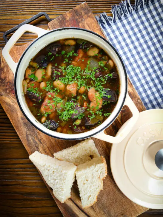 Overhead pork and bean stew with black pudding and sage served in its cooking pot with bread.