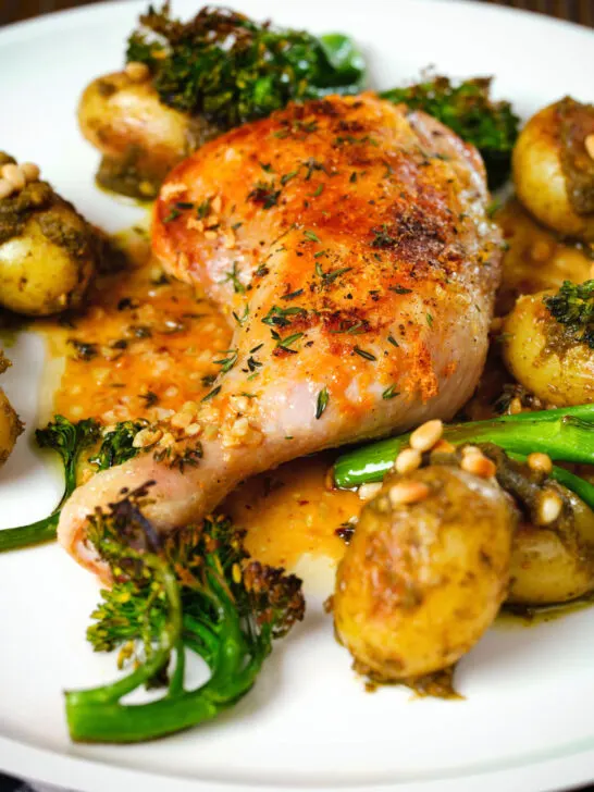 Close-up roasted chicken legs with lemon and thyme served with pesto potatoes and broccoli.